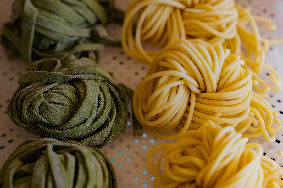 Two coiled bundles of handmade pasta, one green and one yellow, arranged on a perforated white surface.